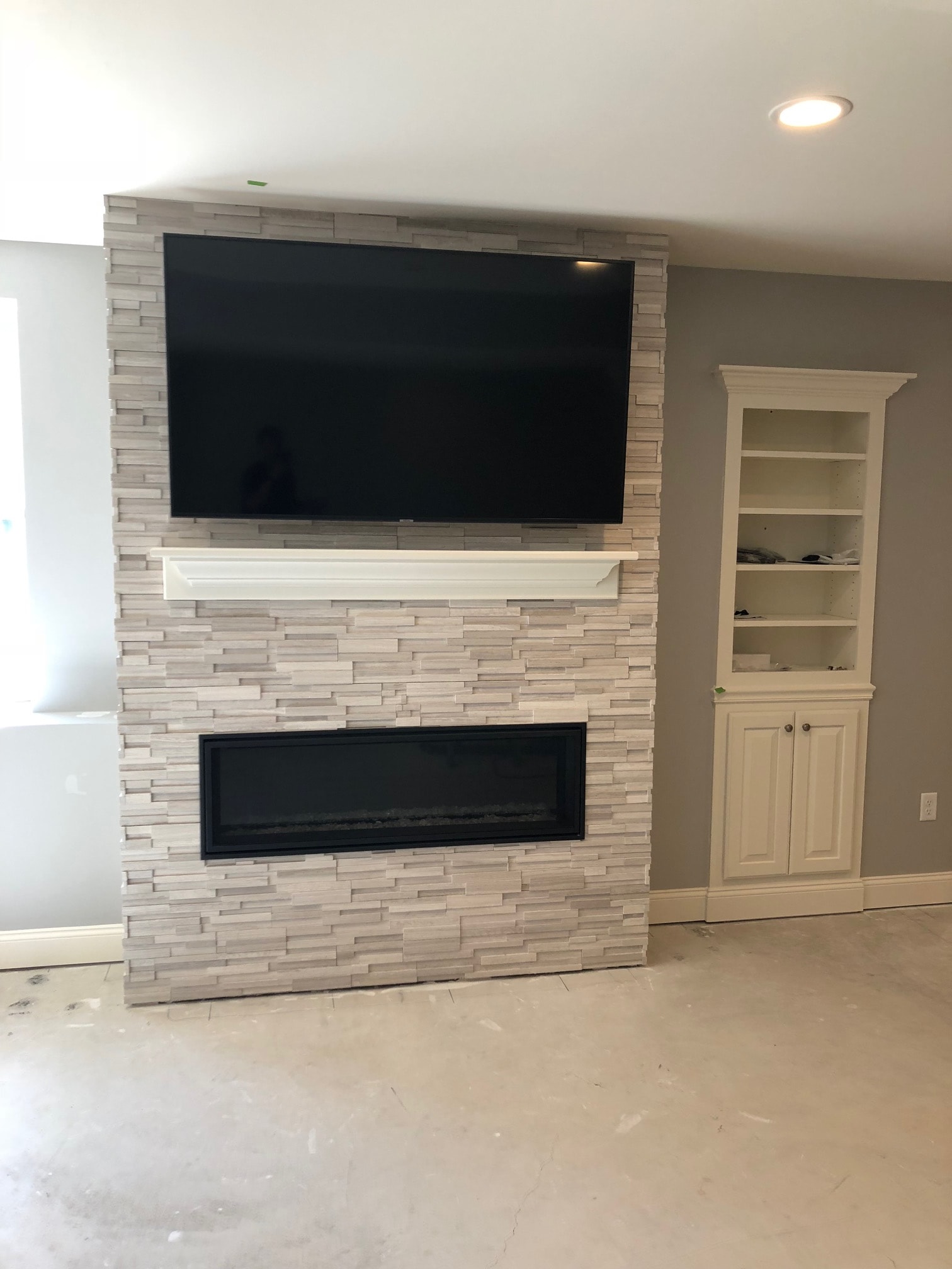 Guilderland basement fireplace feature wall with stacked stone tile surround, mounted TV, black-framed niche, and upper built-in cabinets with panel doors